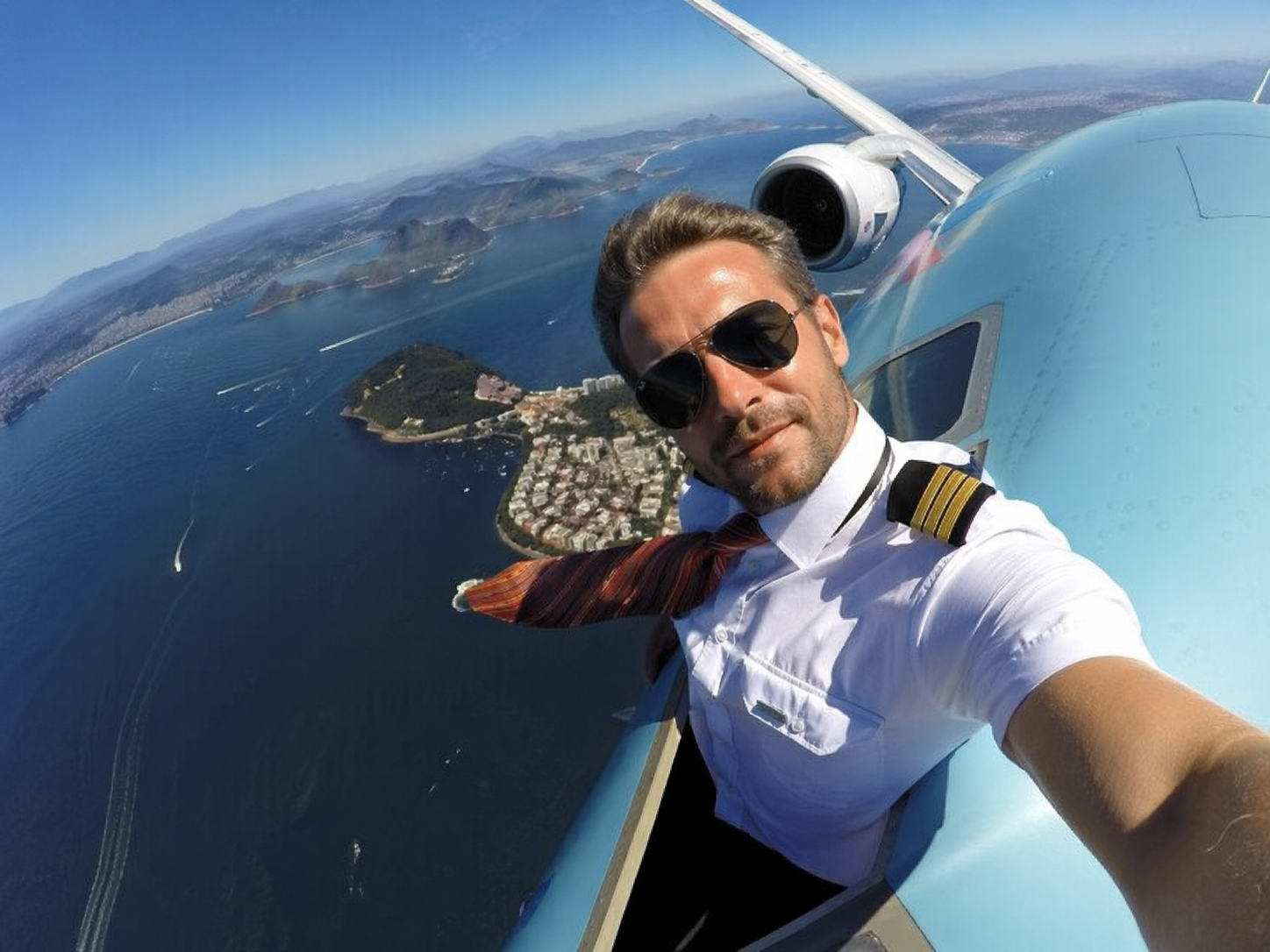 Pilot si dělá selfie venku na letadle za letu, pod ním je vidět křídlo a krajina
